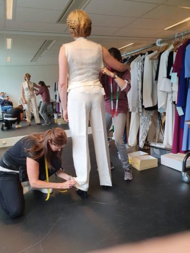 Zwei Schneiderinnen nehmen Maß an einer Frau, die ein weißes Outfit trägt, inmitten von Kleiderständern in einem Atelier.