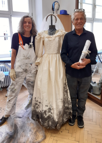 Zwei lächelnde Personen stehen neben einer Schaufensterpuppe mit einem weißen, am Saum verschmutzten Hochzeitskleid in einem hellen Raum.
