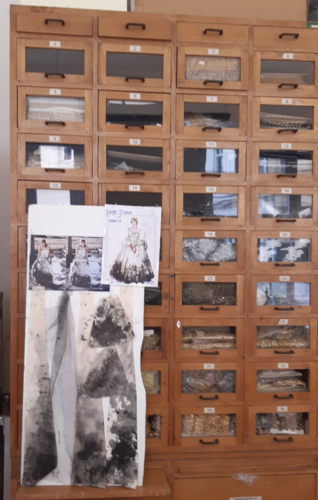 Ein hölzernes Schubladenschrank mit nummerierten Fächern, darauf Modezeichnungen und Stoffproben befestigt.