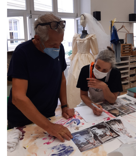 Zwei Personen mit Masken arbeiten an einem Tisch mit Fotos und Kunstmaterialien, im Hintergrund steht ein Kleid auf einer Schneiderpuppe.