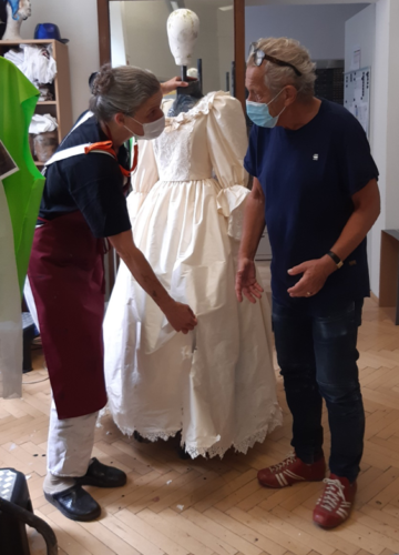Zwei Personen mit Masken besprechen ein weißes historisches Kleid an einer Schneiderpuppe in einem Atelier.