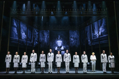 Eine Gruppe von Menschen in einheitlichen Anzügen steht nebeneinander auf einer Theaterbühne vor dunklen Porträts und einem königlichen Bild.