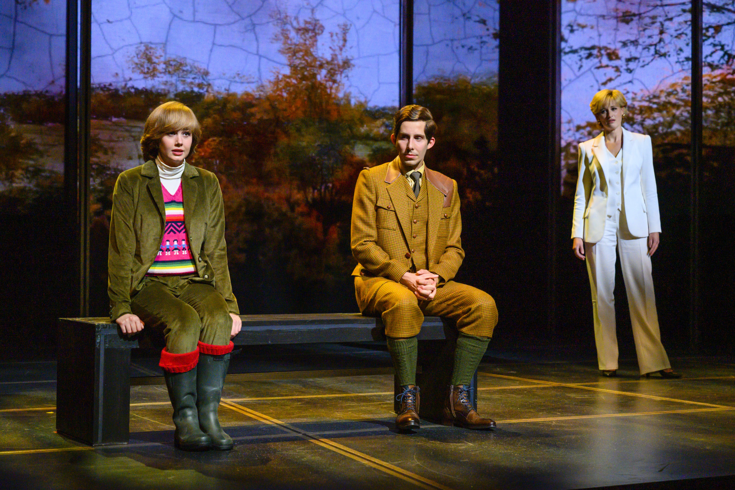 Drei Personen stehen auf einer Theaterbühne; eine sitzt auf einer Bank, zwei stehen im Hintergrund mit herbstlicher Waldkulisse.