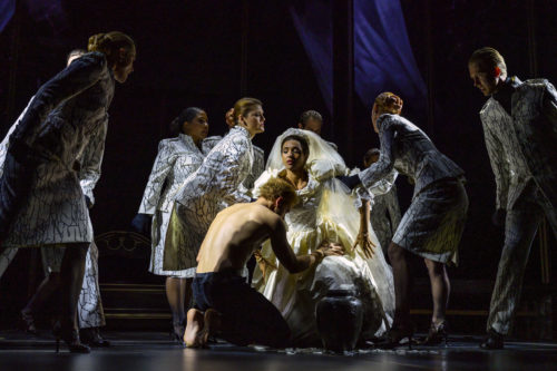 Mehrere Menschen in auffälligen schwarz-weißen Outfits umringen eine Frau im weißen Kleid und Schleier in dramatischer Theaterbeleuchtung.
