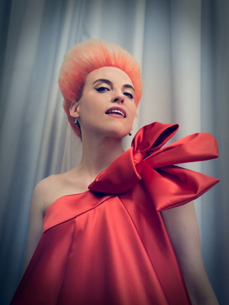 Eine Person mit einer auffälligen rosa Frisur trägt ein schulterfreies rotes Kleid mit großer Schleife vor hellem Vorhang.