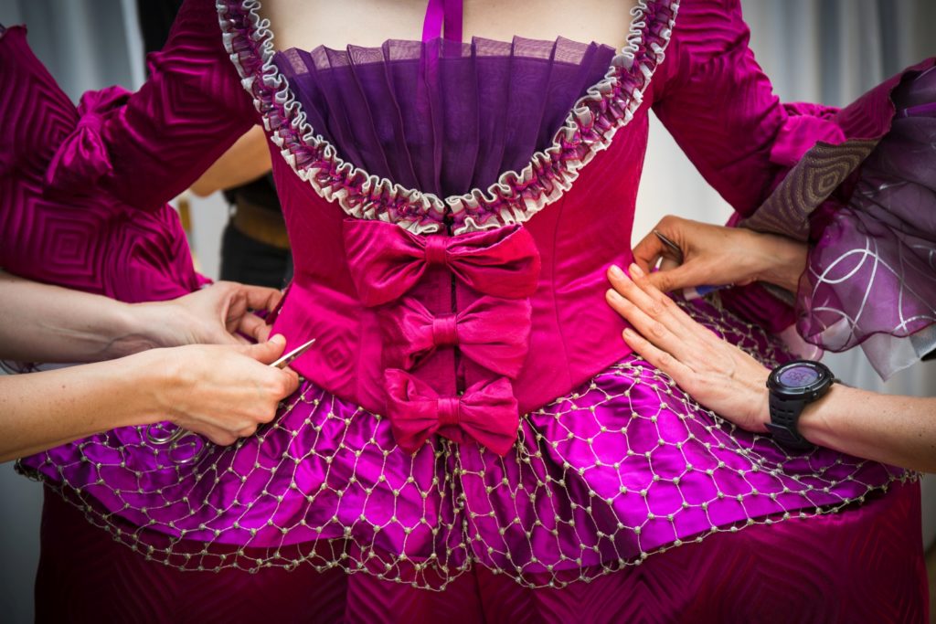 Mehrere Hände helfen dabei, das detailreiche, violett-pinke Bühnenkostüm mit Schleifen und Netz an einer Person zu richten.