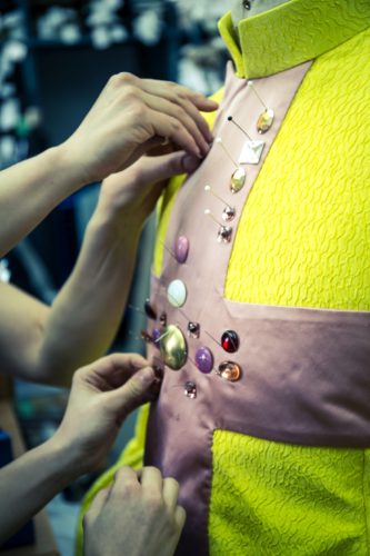 Mehrere Hände stecken bunte Schmuckstücke und Stecknadeln an ein gelbes Stoffkleid mit rosa Einsatz.