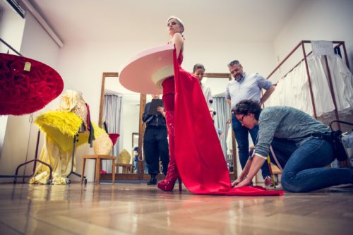 Eine Frau trägt ein auffälliges rotes Kleid mit langem Umhang, während zwei Personen am Rock arbeiten und andere im Hintergrund zuschauen.