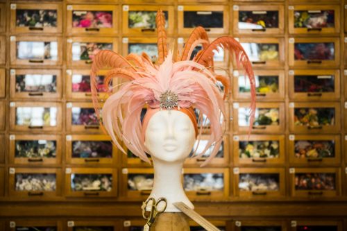 Eine Schaufensterpuppe mit auffälligem rosa Federkopfschmuck steht vor einem Holzregal mit vielen kleinen Fächern.