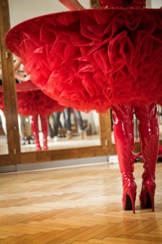 Person in einem roten Tutu-Kleid und glänzenden roten Overkneestiefeln steht vor einem Spiegel auf Parkettboden.