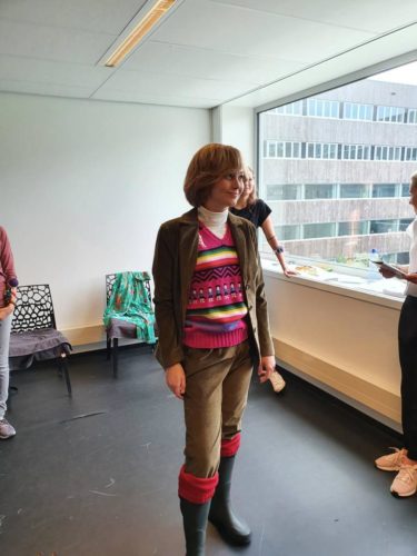Eine Frau mit braunen Haaren trägt einen braunen Anzug mit pinkem Pullover und steht in einem hellen Raum vor einem Fenster.