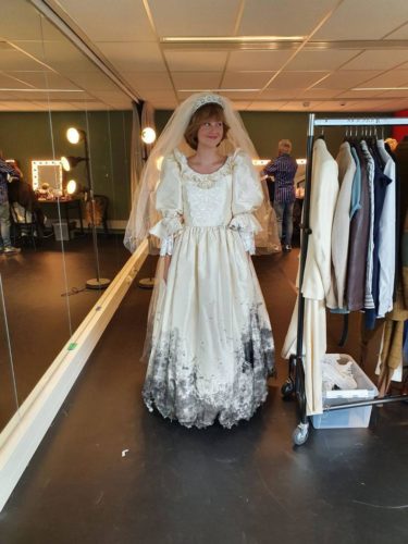 Eine Person trägt ein altes, verschmutztes Brautkleid mit Schleier in einem Umkleideraum mit Spiegeln und Kleiderstange.