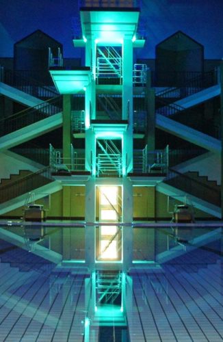 Beleuchteter Sprungturm mit mehreren Plattformen spiegelt sich im Schwimmbecken eines Hallenbads wider.