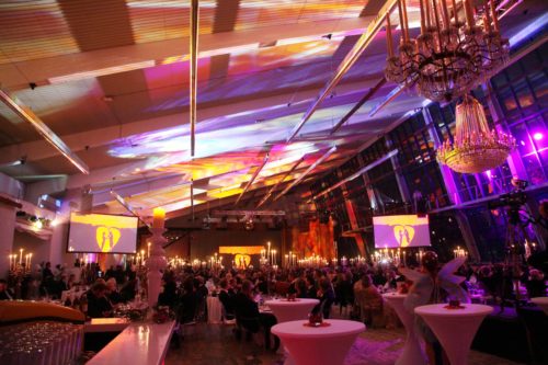 Großer, festlich beleuchteter Ballsaal mit gedeckten Tischen, Kerzen und vielen elegant gekleideten Gästen.