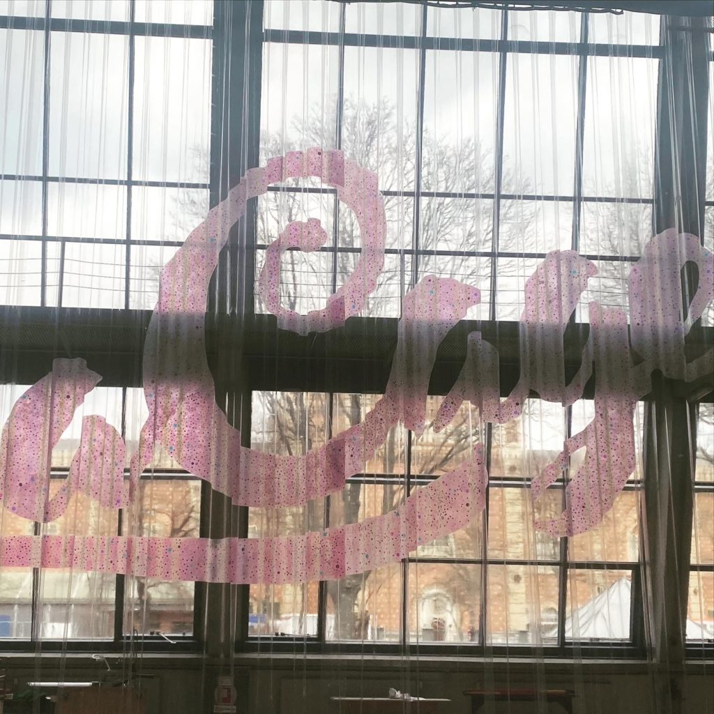 Große, pink gepunktete Aufschrift „Café“ auf einem transparenten Vorhang vor einem großen Fenster mit Blick auf Bäume und Gebäude.