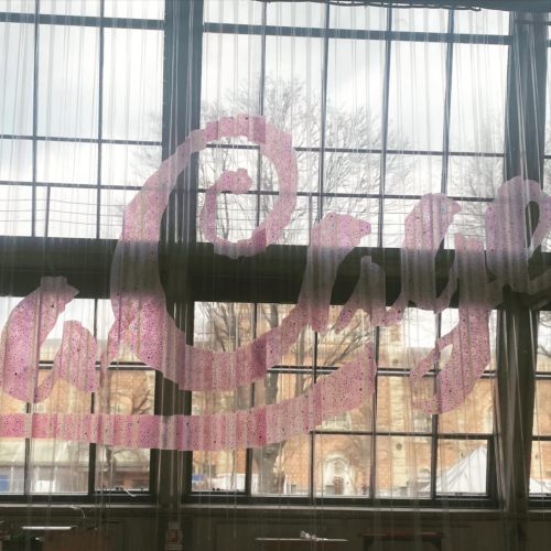 Große, pink gepunktete Aufschrift „Café“ auf einem transparenten Vorhang vor einem großen Fenster mit Blick auf Bäume und Gebäude.