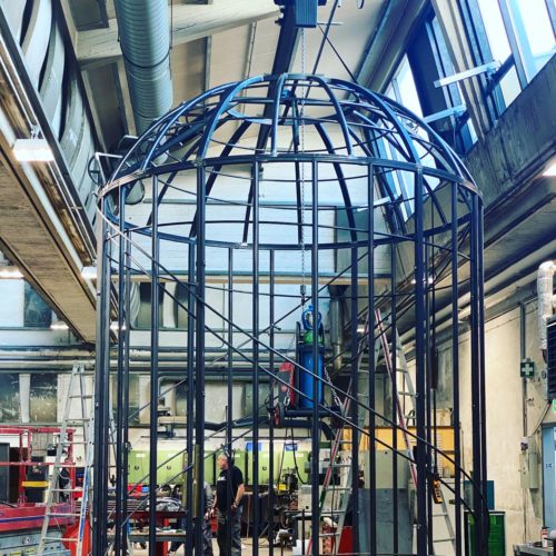 Eine große Metallkuppel wird in einer Werkstatt aufgebaut, während mehrere Personen daran arbeiten.