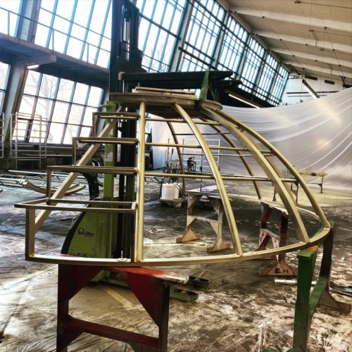 Metallgerüst in Kuppelform steht in einer großen Werkhalle mit hohen Fenstern und wird vermutlich bearbeitet oder montiert.