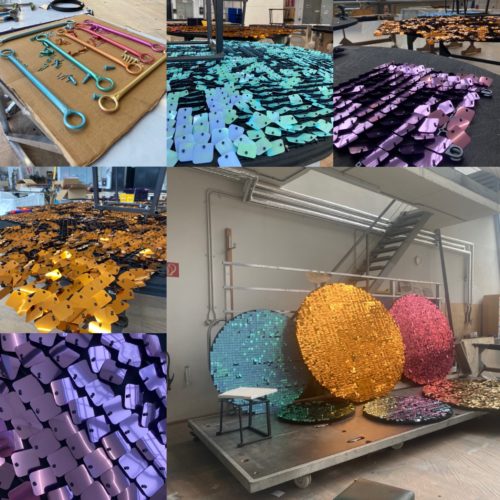 Bunte, glänzende Metallplättchen in Blau, Lila, Orange und Gold liegen gestapelt und wurden zu großen, kreisförmigen Kunstobjekten angeordnet.