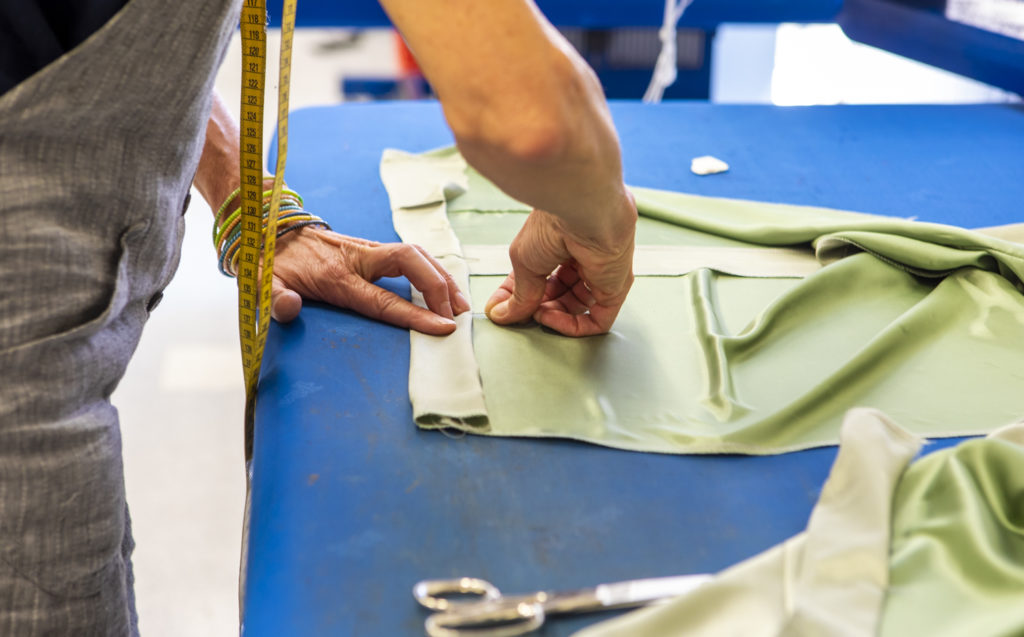 Eine Person arbeitet mit grünem Stoff auf einem blauen Tisch, daneben liegt eine Schere und ein Maßband.
