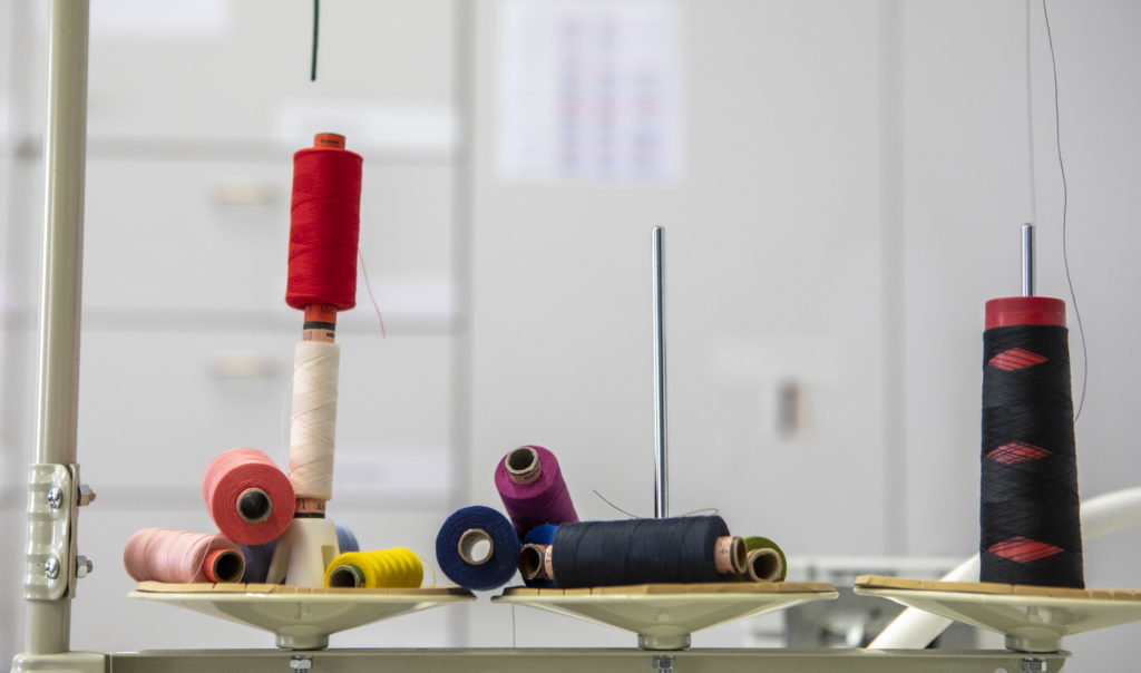 Verschiedene bunte Garnrollen stehen und liegen nebeneinander auf weißen Haltern in einer Werkstatt oder Nähumgebung.