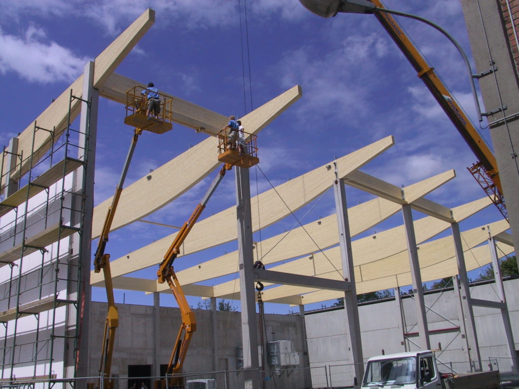Zwei Bauarbeiter auf Hebebühnen montieren große Holzbalken auf einer Baustelle eines Gebäudes bei sonnigem Wetter.