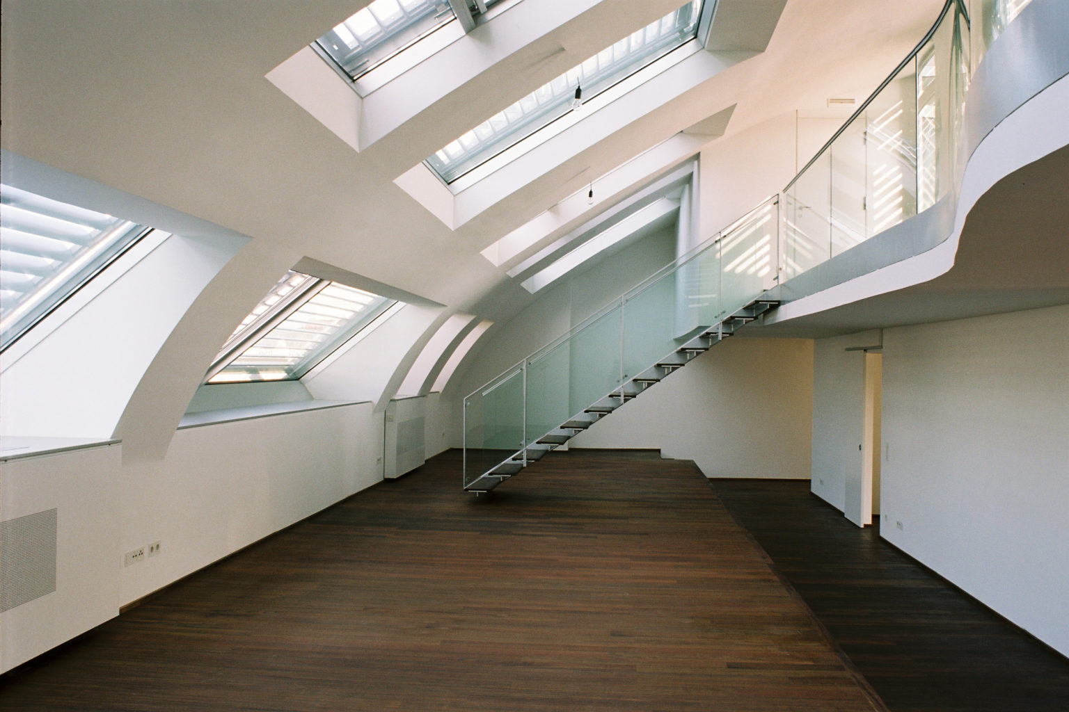 Großer, moderner Dachgeschossraum mit Holzboden, gewölbten Fenstern und einer Treppe mit Glasgeländer.