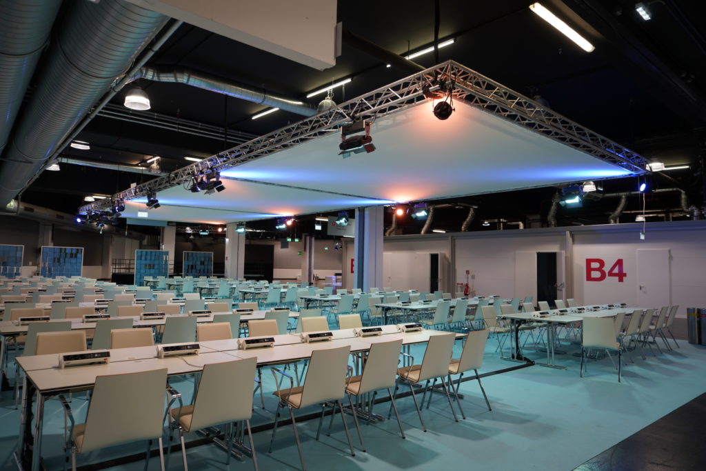 Großer Konferenzraum mit vielen leeren weißen Stühlen und Tischen, moderne Beleuchtung und sichtbarem Belüftungssystem, Bereich B4.