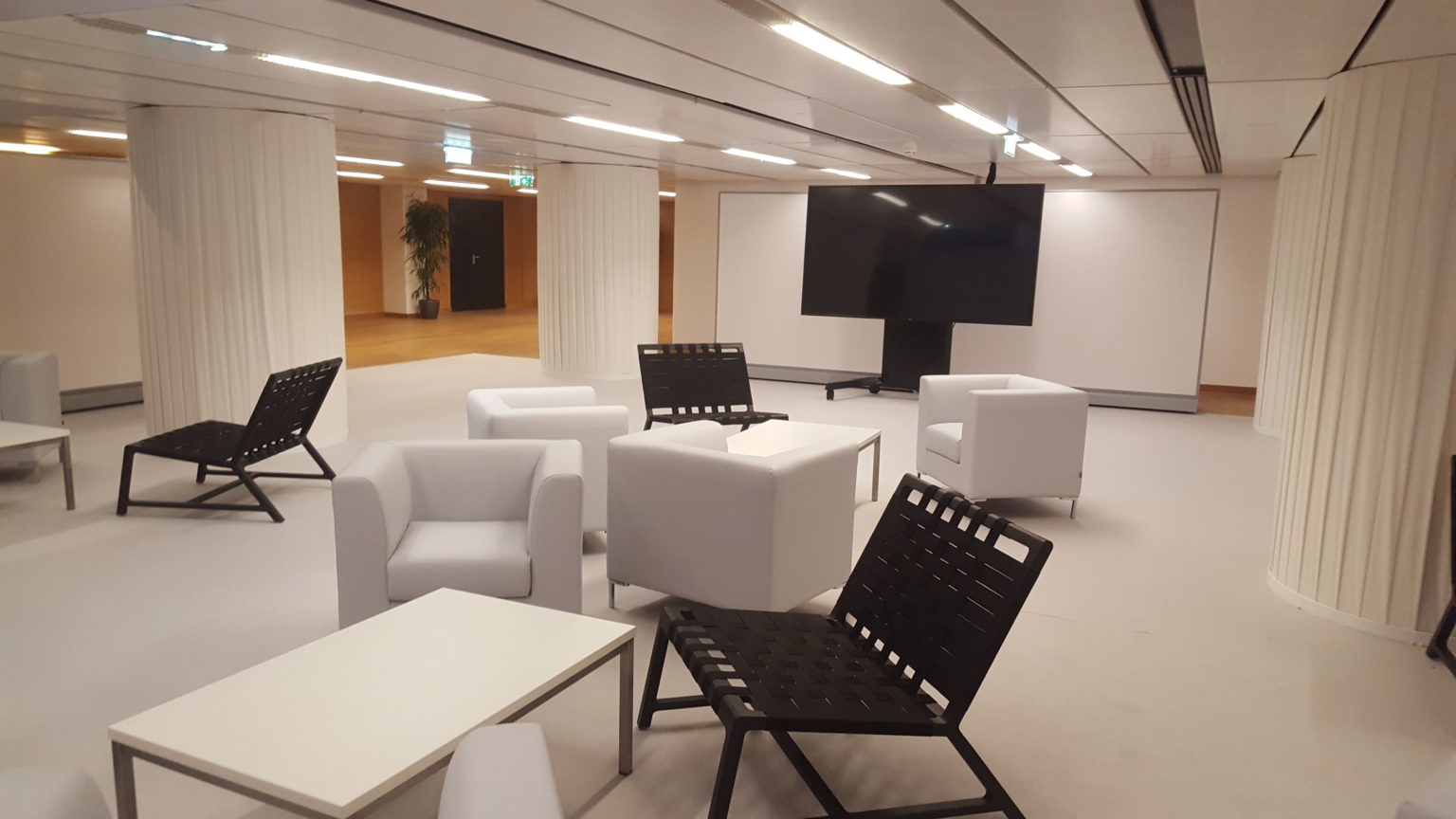 Moderne Lounge mit weißen Sesseln, schwarzen Stühlen, Couchtischen und einem großen Fernseher in einem hellen Raum.