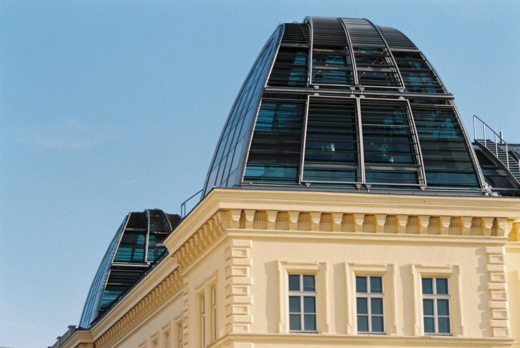 Moderne gläserne Kuppeln auf einem klassischen hellgelben Gebäude mit mehreren Fenstern vor blauem Himmel.