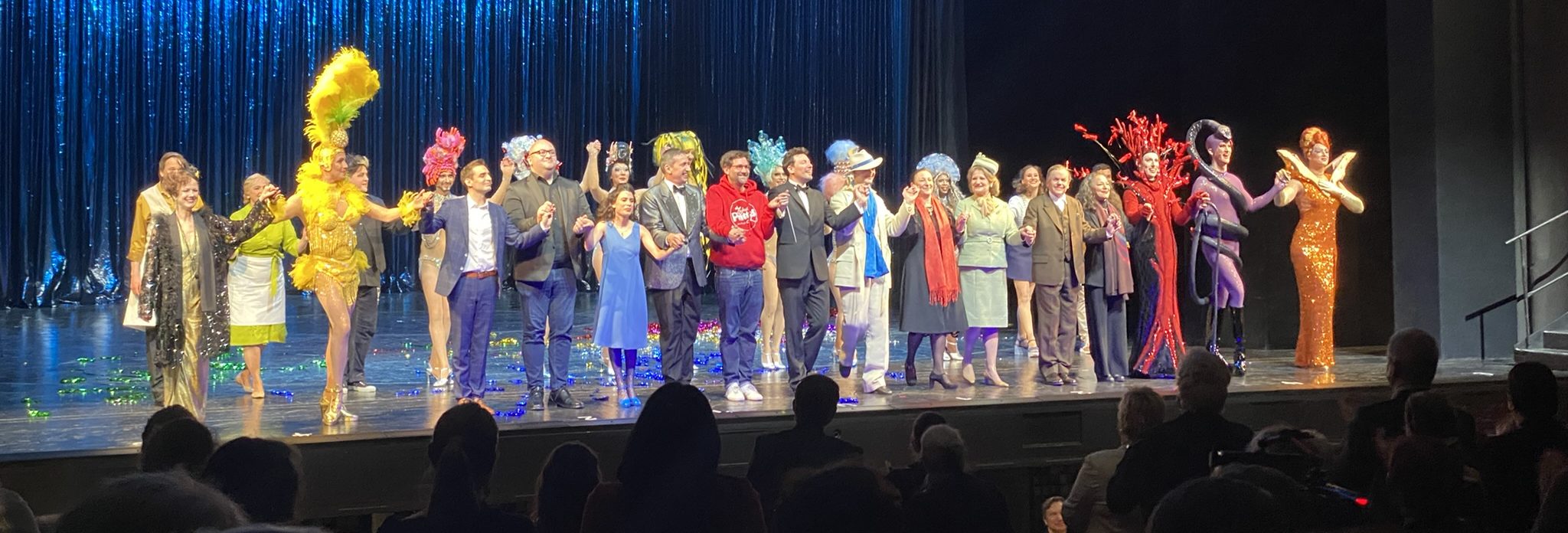 Ensemble von Bühnenkünstlern in farbenfrohen Kostümen steht auf einer Theaterbühne und hält gemeinsam die Hände zum Applaus.