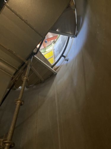 Blick von innen durch eine runde Öffnung in einem großen Metallbehälter mit sichtbaren Gerüstteilen und Tageslicht am oberen Rand.