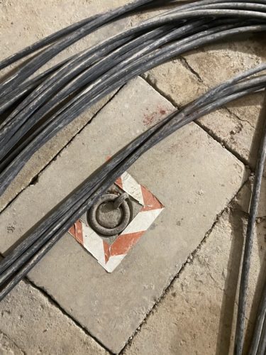 Mehrere schwarze Kabel verlaufen über einen Boden aus Steinfliesen, in deren Mitte eine markierte Fliese mit Metallring eingelassen ist.