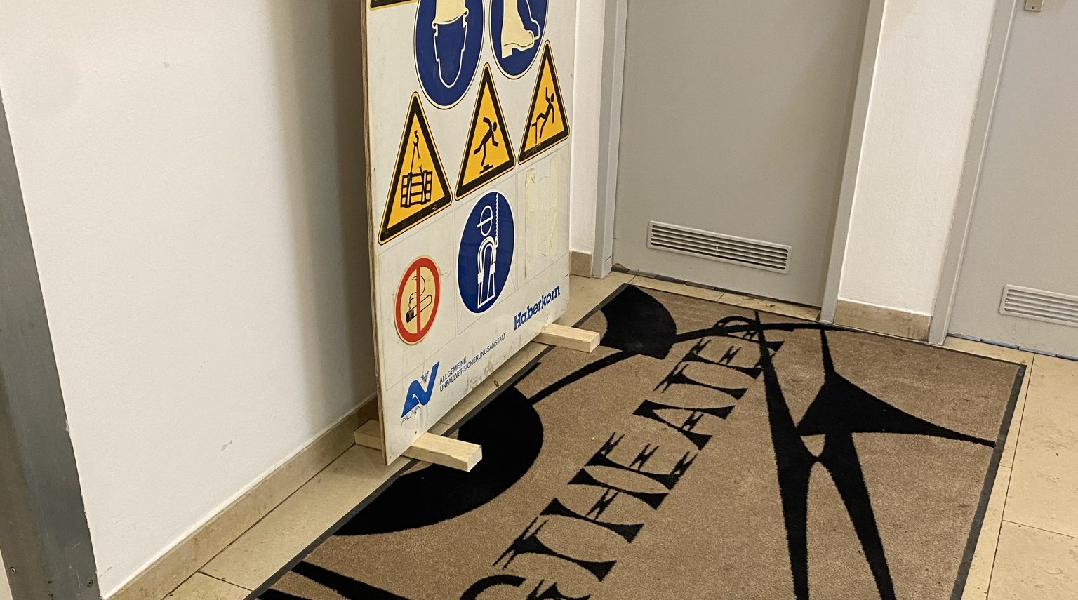 Ein Schild mit verschiedenen Sicherheits- und Warnsymbolen steht auf einem Flur neben einer Tür auf einer Matte mit „THEATER“-Schriftzug.