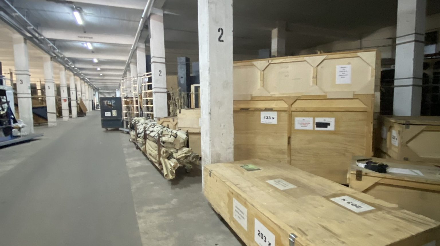 Große Lagerhalle mit nummerierten Säulen, Holzkisten und Kisten am rechten Rand, einige mit sichtbaren Beschriftungen.
