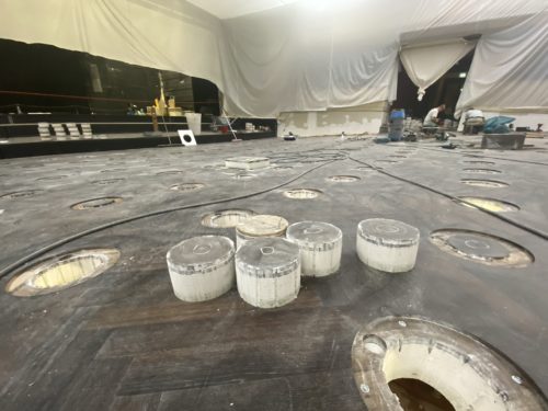 Eine Baustelle mit einer hölzernen Bodenfläche, in die zahlreiche runde Ausschnitte und mehrere durchsichtige Kunststoffzylinder eingesetzt sind.