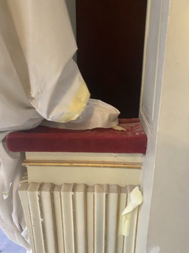 Ein rotes Samtpolster liegt auf einer hellen Heizkörperabdeckung, darauf und daneben liegen abgedeckte, mit Klebeband fixierte Tücher.