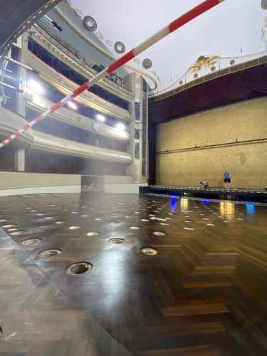 Blick auf eine Theaterbühne mit vielen runden Öffnungen im Boden, zwei Personen arbeiten im Hintergrund, roter Absperrbalken oben.