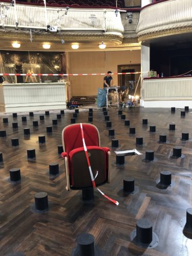 Ein einzelner roter Theatersessel steht abgesperrt in einem Saal mit vielen schwarzen Podesten auf dem Boden, im Hintergrund Bauarbeiten.