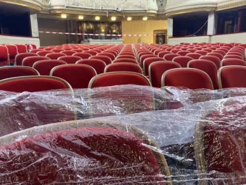 Reihen von roten Theatersesseln, einige mit Plastikfolie abgedeckt, und Absperrband im Hintergrund.
