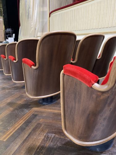 Reihe von Theatersitzen mit Holzverkleidung und roten Samtpolstern auf einer Bühne oder in einem Theatersaal.
