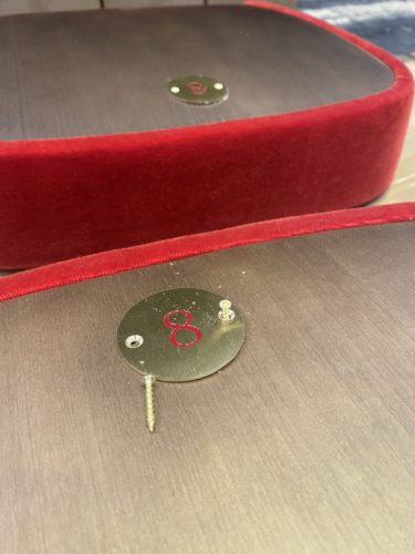 Eine rot umrandete Holzbasis mit einem runden Metallschild, das eine rote Acht sowie eine Schraube und Dübel zeigt.