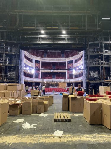 Bühnenansicht eines Theaters mit vielen Kartons, leeren Stühlen und mehreren Menschen beim Aufbau.