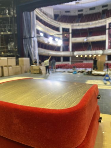 Rote Stuhlkissen im Vordergrund einer Theaterbühne mit Menschen und Scheinwerfern bei Bühnenarbeiten im Hintergrund.