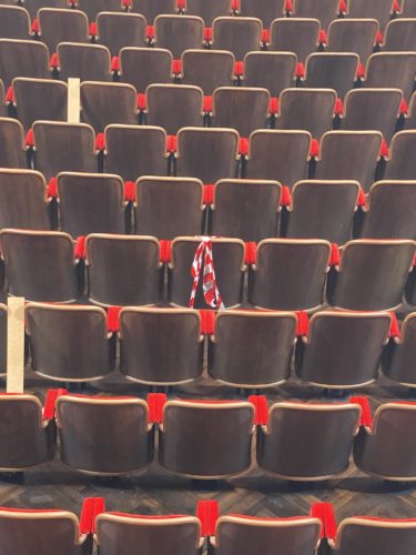 Mehrere leere Sitzreihen in einem Saal, wobei ein Sitz mit rot-weißem Flatterband abgesperrt ist.