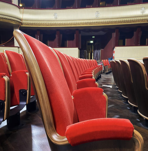 Eine Reihe roter Theatersessel in einem eleganten, leeren Saal mit goldener Balustrade.