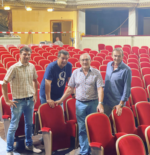 Vier Männer stehen in einem Theater zwischen roten Sitzreihen und lächeln in die Kamera.
