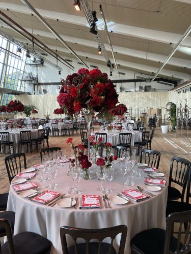 Festlich gedeckter runder Tisch mit weißer Tischdecke, roten Blumengestecken und elegant arrangiertem Besteck in einem großen Saal.