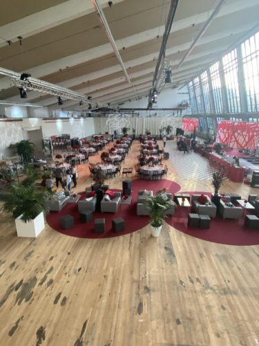 Großer, modern dekorierter Veranstaltungssaal mit gedeckten runden Tischen, Lounge-Bereichen und großen Fenstern.