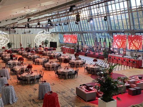 Großer, elegant dekorierter Veranstaltungssaal mit runden Tischen, roten Blumenarrangements und Bühne für Live-Musik.